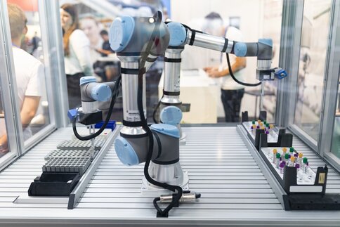 Ein automatisierter, blau-grauer Roboterarm im Laborumfeld auf der Messe analytica. Er ist in einer Arbeitsstation platziert, die mit Probenhaltern ausgestattet ist. Im Hintergrund sind unscharf einige Personen zu sehen.