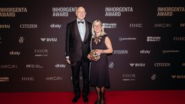 A man and a woman in festive attire stand smiling in front of a photo wall of the INHORGENTA AWARD, holding a trophy.
