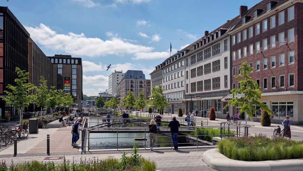  A lively city square with water features, green recreational areas, and surrounding modern and historic buildings, where people gather on sunny days.