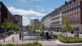  A lively city square with water features, green recreational areas, and surrounding modern and historic buildings, where people gather on sunny days.