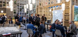 Menschen in Geschäftskleidung sitzen in einem Messestand der aus einer komplexen Holzkonstruktion besteht, mit stimmungsvollen Hängelampen.