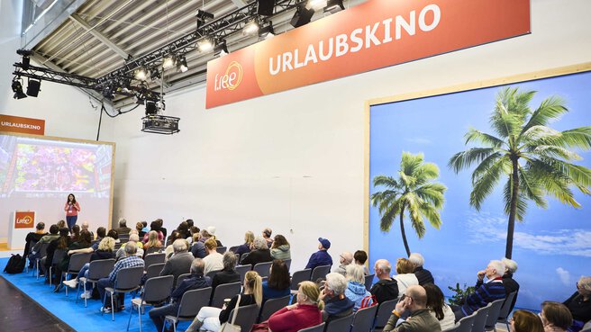 Ein Vortrag im „Urlaubskino“ auf der Reisemesse f.re.e, mit Publikum, Leinwand und tropischem Palmenbild im Hintergrund.