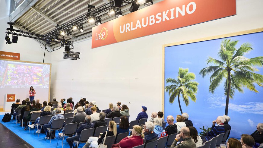 A lecture in the “vacation cinema” at the f.re.e travel trade fair, with an audience, screen and tropical palm trees in the background.