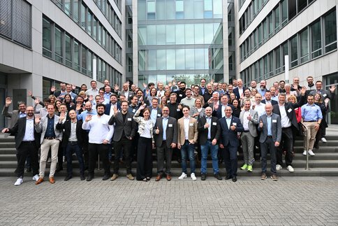 Eine große Gruppe von Menschen posiert und lächelt auf den Stufen vor einem modernen Bürogebäude.