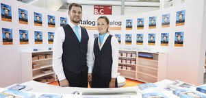 Two trade fair employees at a stand with several catalog displays on the walls and a counter.