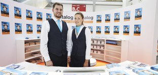 Two trade fair employees at a stand with several catalog displays on the walls and a counter.