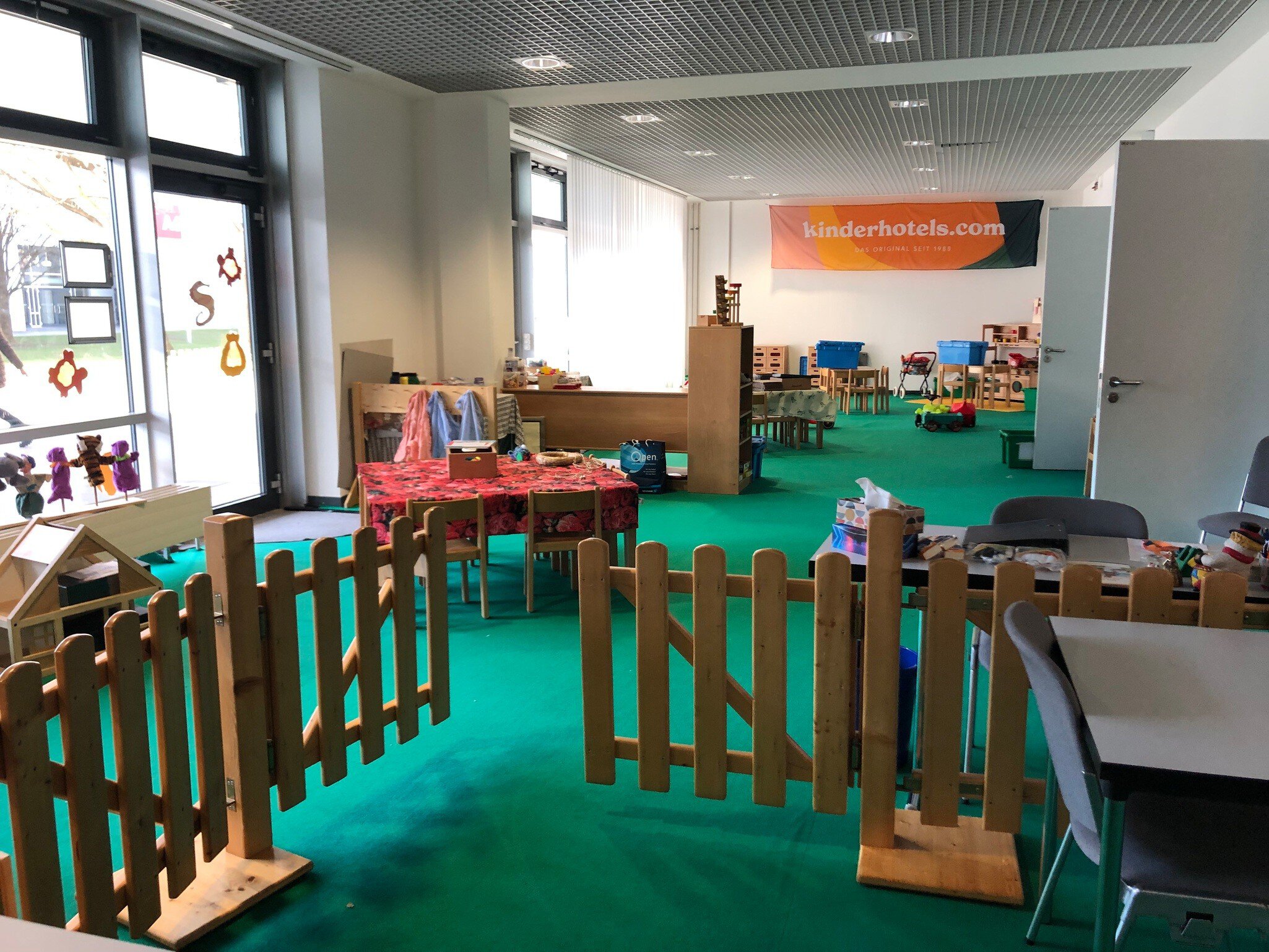 Ein großer Raum mit grünem Teppichboden, Kinderspielzeug und niedrigen Tischen. Im Hintergrund hängt ein Banner für „kinderhotels.com“.