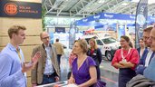 Young woman in a purple dress attentively listens to a young man in a light blue shirt, while five other people (also Bruno Rudnik) follow the presentation at the „Solar Materials“ booth as well.
