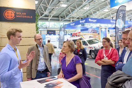 Young woman in a purple dress attentively listens to a young man in a light blue shirt, while five other people (also Bruno Rudnik) follow the presentation at the „Solar Materials“ booth as well.