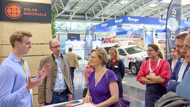Young woman in a purple dress attentively listens to a young man in a light blue shirt, while five other people (also Bruno Rudnik) follow the presentation at the „Solar Materials“ booth as well.