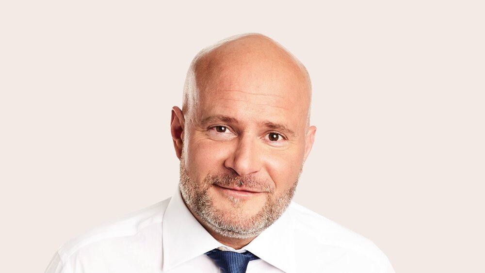 Glatzköpfiger Mann mit Bart, weißem Hemd und blauer Krawatte, lächelt vor hellem Hintergrund in die Kamera.