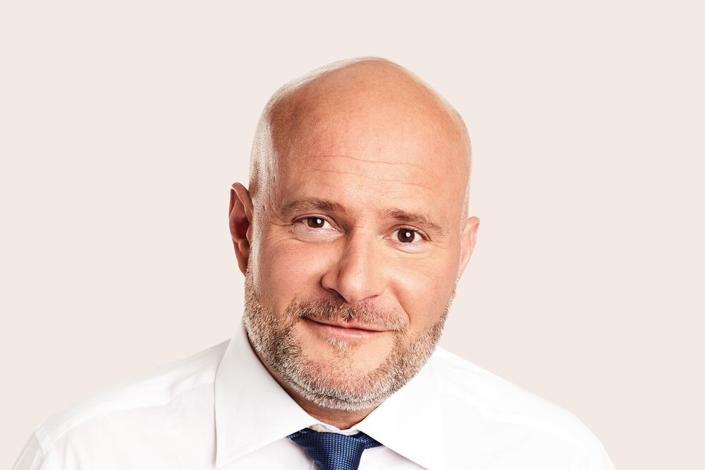 Glatzköpfiger Mann mit Bart, weißem Hemd und blauer Krawatte, lächelt vor hellem Hintergrund in die Kamera.