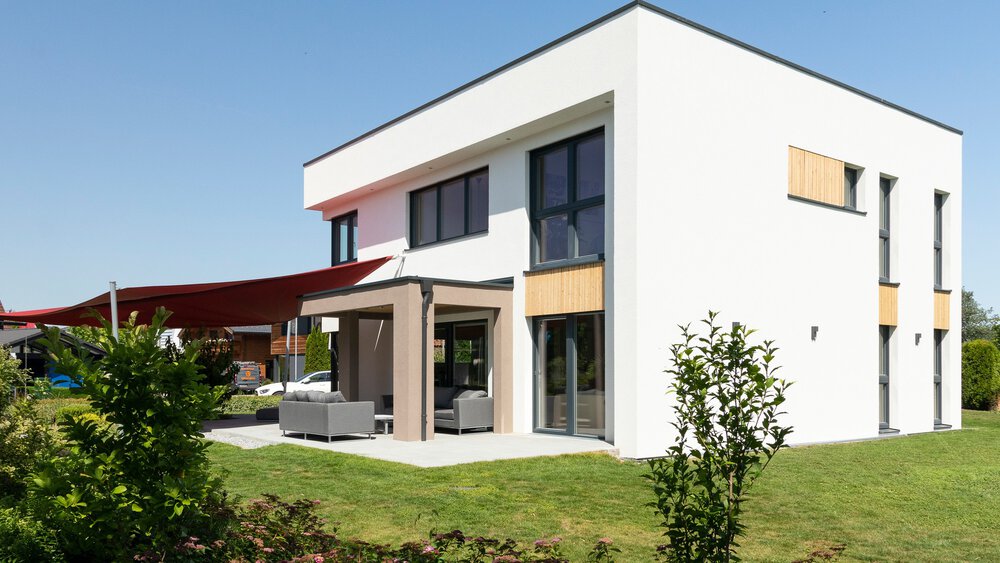 Modernes weißes zweistöckiges Haus mit großen Fenstern, Terrasse, Gartenmöbeln und grünem Rasen unter strahlend blauem Himmel.