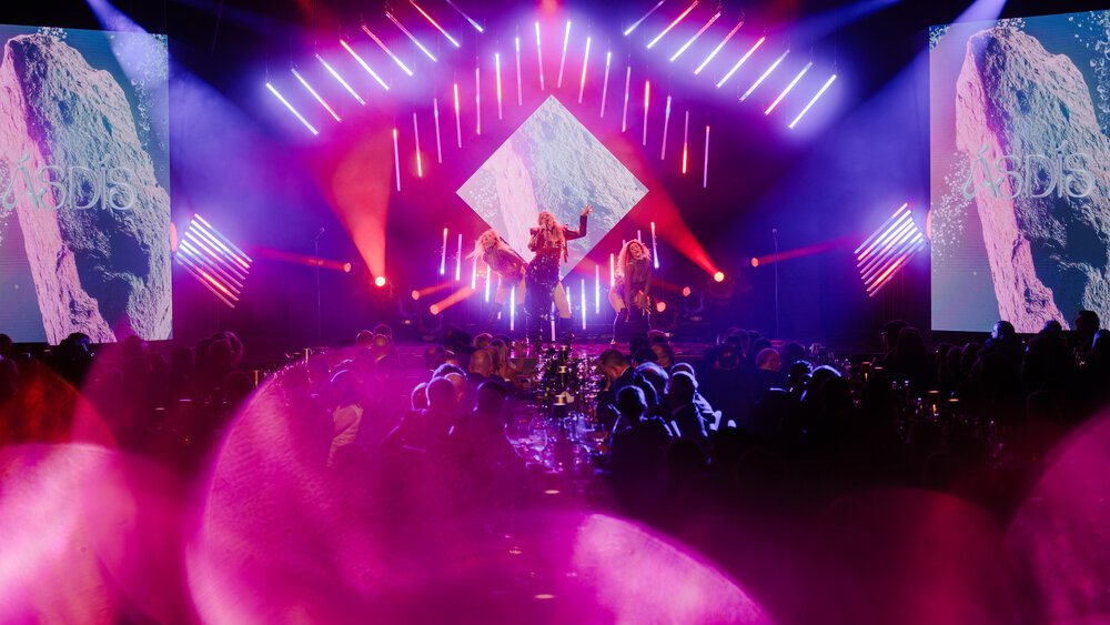 Konzertbühne mit leuchtend rosa und lila Lichtern, Darstellern und großen Bildschirmen, davor ein Publikum.