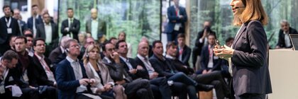 Eine Frau im Anzug spricht zu einem Publikum aus Fachleuten in einem modernen Konferenzraum.