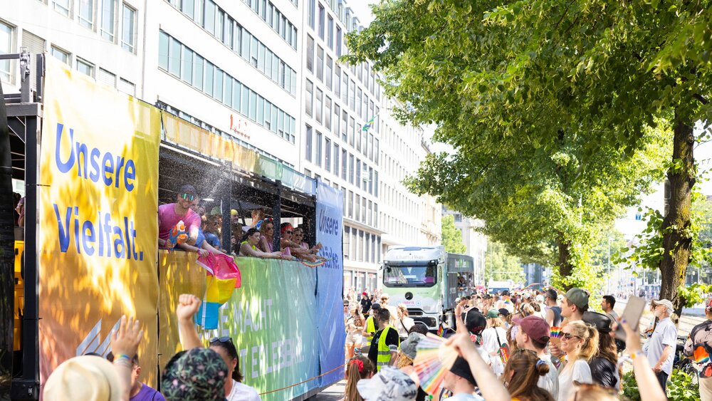People are celebrating at a parade featuring a float bearing the words “Our Diversity” on a sunny street lined with buildings and trees.