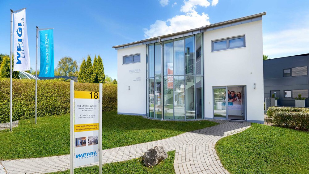 Modernes zweistöckiges Gebäude in Glas- und Weißtönen mit einem Weg und Rasen, klarem blauen Himmel und einem Hinweisschild im Vordergrund.