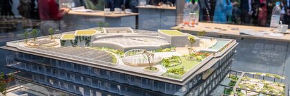 Multi-storey architectural model of a modern building with solar panels and a green roof; trade fair visitors can be seen in the background.