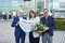 Two men and a woman in business attire stand in front of the Messe München entrance with a bouquet of flowers and a gingerbread heart.