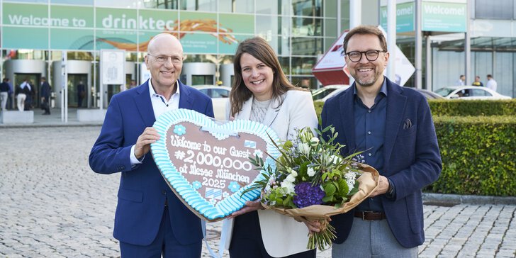 Zwei Männer und eine Frau in Businesskleidung stehen mit einem Blumenstrauß und einem Lebkuchenherz vor dem Messe München Eingang.