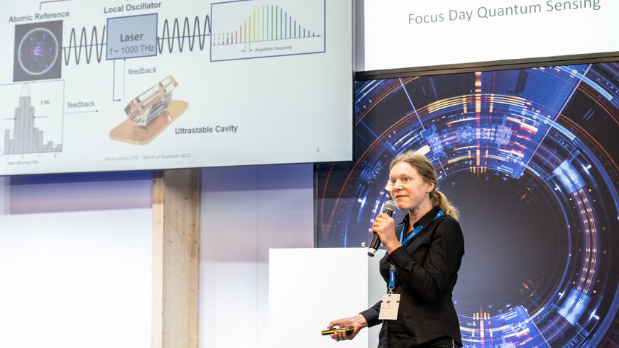 A woman with a microphone is giving a lecture on quantum sensor technology and is standing next to a large display with diagrams and text.
