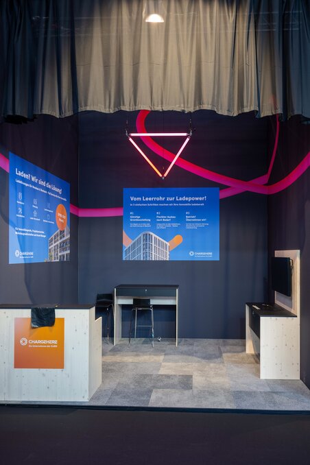 Ein unbesetzter Eckstand auf der EXPO REAL mit Sitzmöglichkeiten, Werbebannern, einer dreieckigen Beleuchtung und schwarzen Vorhängen.