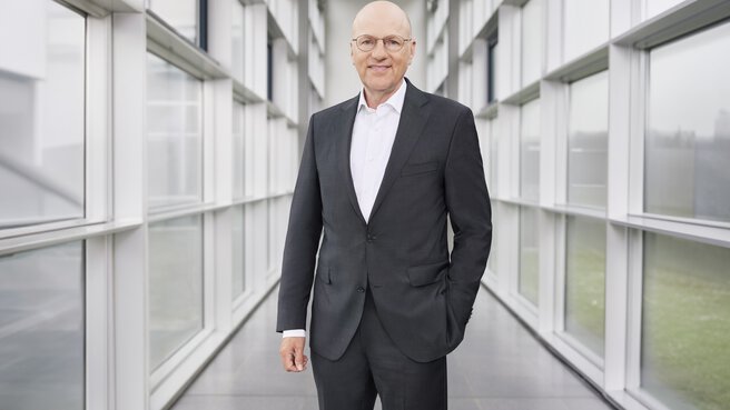 Ein lächelnder Mann im Anzug, mit Glatze und Brille steht mit einer Hand in der Tasche in einem Glaskorridor.