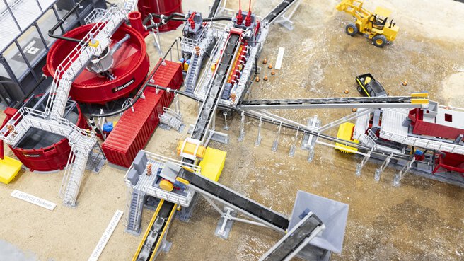 Model of an industrial plant with conveyors, containers, and construction vehicles on a sandy surface.