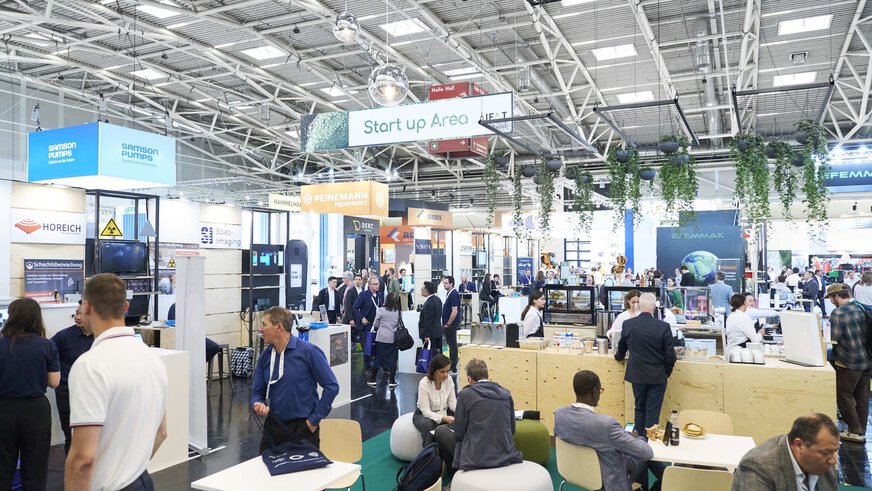 Menschen besuchen die Start up Area auf der IFAT. Im Vordergrund ist eine Sitzecke mit einer Theke, hinten ist eine Reihe an Messeständen zu sehen.