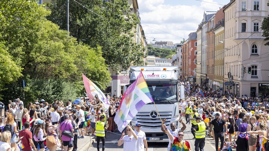 Eine überfüllte Straßenparade mit Menschen, die Regenbogenflaggen halten, und einem Lastwagen in der Mitte.