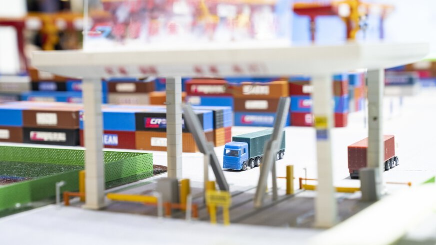 Miniaturdarstellung einer beschrankten Zufahrt zu einem Containerhafen mit LKWs und gestapelten Containern im Hintergrund.