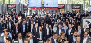 Eine Menschenmenge professionell gekleideter Menschen strömt durch die Eingangshalle der Messe München und geht durch die Ticketschranken.