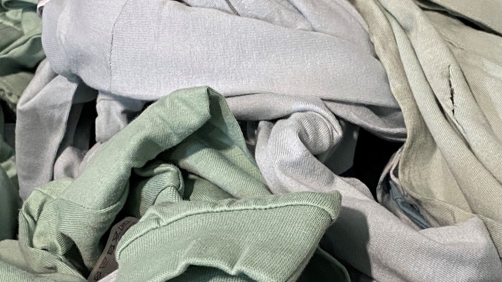 Ein Haufen aus hellgrünen und grauen Jersey‑Stoffen und Kleidungsstücken, locker übereinandergelegt.