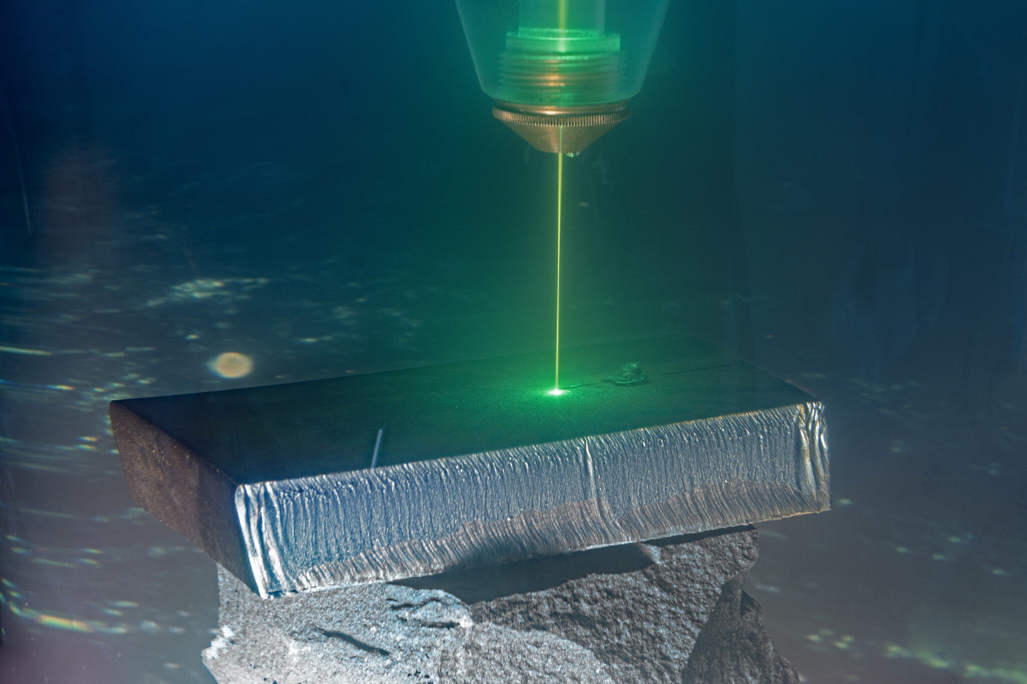 A green laser beam cuts into a metal block standing on a rocky surface under water.