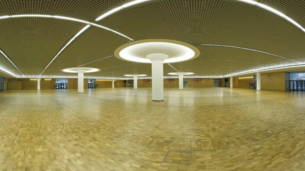 Geräumige, leere Halle mit Holzboden, runden Deckenleuchten und Säulen. An den Wänden befinden sich Rasterdeckenplatten und große Fenster.