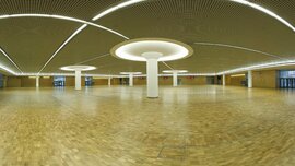 Geräumige, leere Halle mit Holzboden, runden Deckenleuchten und Säulen. An den Wänden befinden sich Rasterdeckenplatten und große Fenster.