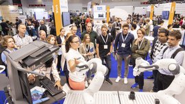 A woman presents the functions of two modern robot arms to an interested audience at the automatica exhibition.