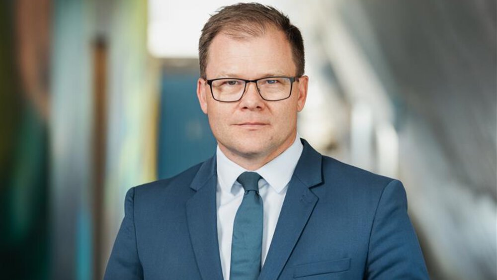 Porträt von Bundesumweltminister Carsten Schneider im blauen Anzug mit Krawatte und Brille, blickt in die Kamera; unscharfer Hintergrund in einem modernen Gebäude.