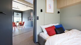 Modernes Schlafzimmer mit einem Bett und bunten Kissen, offene Tür zeigt einen Essbereich mit roten Stühlen und großen Fenstern.