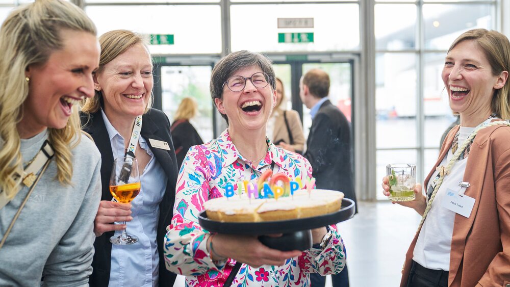 Four women laughing together at the trade fair; one holds a birthday cake with 