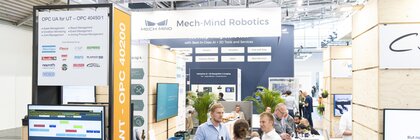 Exhibition booth with the sign “Connected Machines” in a brightly lit trade fair hall. In the foreground, an information desk with monitors and logos of companies such as Atlas Copco and Bosch Rexroth is visible. In the background, Mech-Mind Robotics showcases AI-powered automation solutions. Visitors are engaged in conversation.