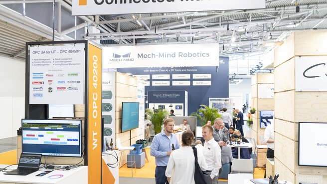 Exhibition booth with the sign “Connected Machines” in a brightly lit trade fair hall. In the foreground, an information desk with monitors and logos of companies such as Atlas Copco and Bosch Rexroth is visible. In the background, Mech-Mind Robotics showcases AI-powered automation solutions. Visitors are engaged in conversation.