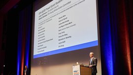 On the stage of the World of Photonics Congress, a man at the podium presents the award winners displayed on the screen.