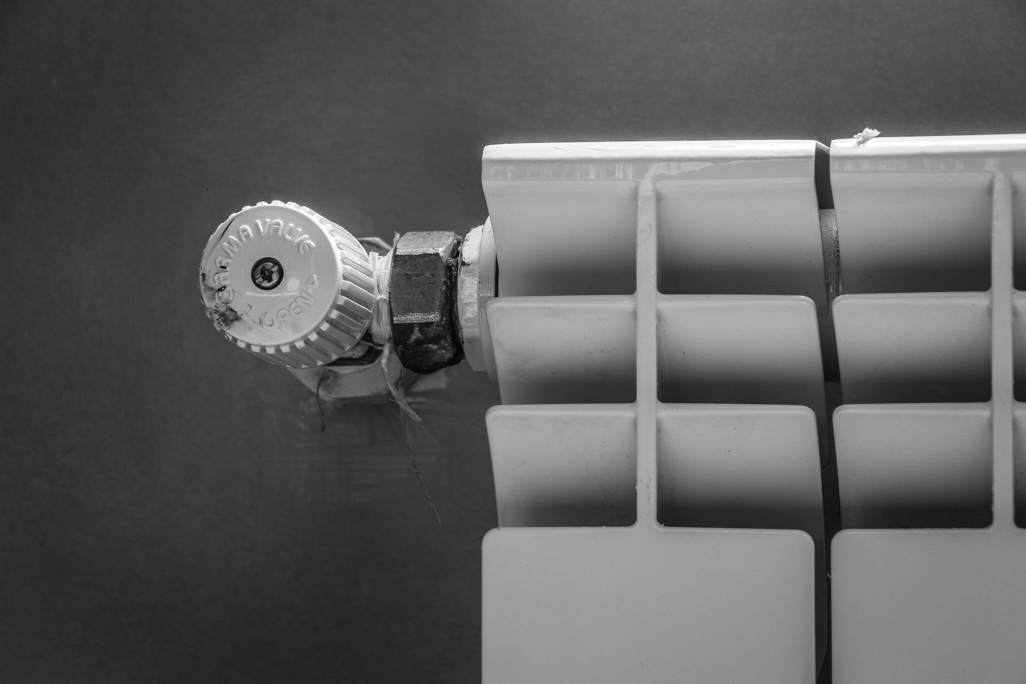 A black-and-white close-up image of a white radiator with fins, a radiator thermostat on the left-hand side and a grey wall behind it.