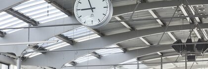 Große Wanduhr mit schlichtem Design, die von einer hohen Decke in einem modernen, gut beleuchteten Raum hängt.