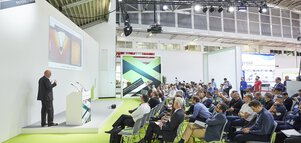 A speaker presents on stage at the Laser Materials Processing Photonics Forum in an exhibition hall.