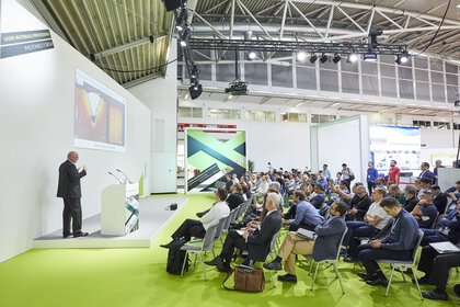 Ein Redner präsentiert auf der Bühne des Laser Materials Processing Photonics Forum in einer Messehalle.