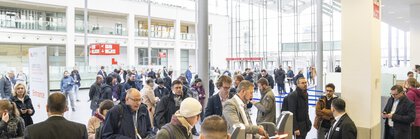 Menschen gehen durch die Ticketschranken in einem belebten, modernen Atrium mit Glaswänden und -decken.
