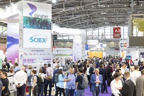 Crowded exhibition hall with large booths from companies such as Danaher and Sciex, many attendees on a purple carpet, gathering information about exhibited laboratory and analytical technology.