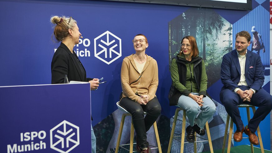 Three people on stools discussed with a moderator at a lectern on a stage at ISPO Munich.